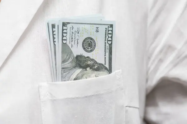 A stack of hundred dollar bills in the pocket of a doctor\'s coat.  Concept: bribe to doctor, corruption in medicine.