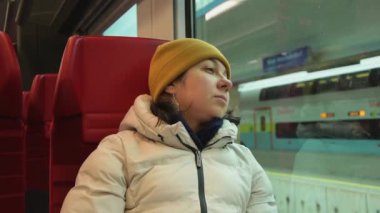 Bir banliyö elektrikli treninin modern bir vagonunda oturan kadın, pencereden dışarı bakıyor, bir Avrupa tren istasyonunun platformu. Konsept: seyahat ve turizm, aile ilişkileri.