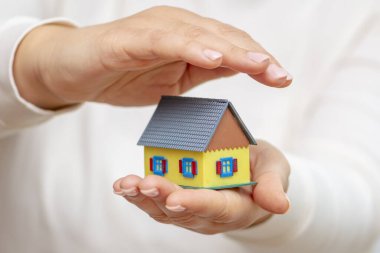 Bir kadın, avucunun içinde minyatür bir ev modelini dikkatlice tutuyor. Diğer eli ise sevgiyle korunuyor ve kırılgan yapıyı koruyor..