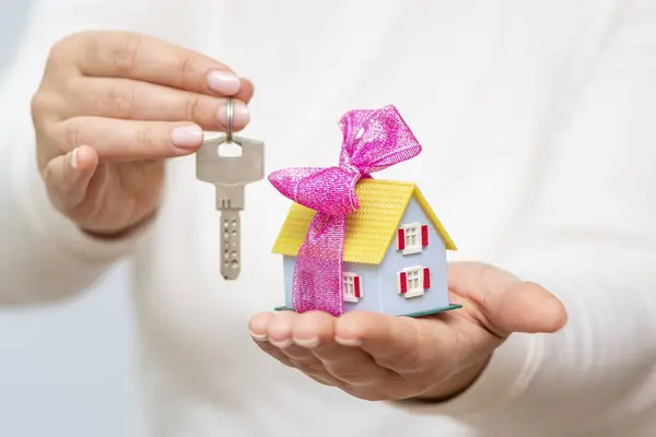 The woman is holding a mini house with a bow and metal keys to the apartment.