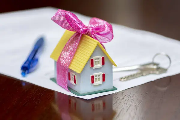 A model of a mini house with a bow, apartment keys, and a sheet of paper lying on a table. Concept: buying and selling housing.