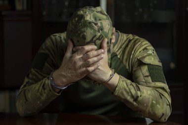Kamuflaj üniformalı asker. Kapalı yerde kelepçeli. Yüzü koyu renk..
