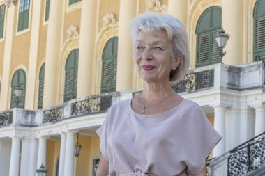 Yaşlı bir kadın parlak sarı bir binanın önünde duruyor ve gülümsüyor..