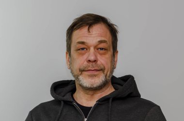 50-55 yaşlarında, sakallı, siyah kapüşonlu yaşlı bir adam açık renkli bir arka planda kameraya bakıyor..