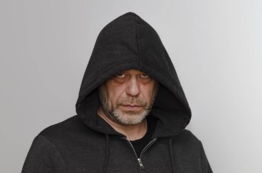 50-55 yaşlarında, sakallı, siyah kapüşonlu kapüşonlu giyen yaşlı bir adam gri bir arka planda kameraya bakıyor..