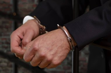 Takım elbiseli bir adam kelepçeli, elleri parmaklıklarda dinleniyor, tuğla dövmenin bulanık arkaplanı...