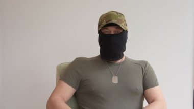 Askeri üniformalı ve şapkalı bir adamın portresi, siyah kar maskesi takıyor, kameraya bakıyor ve konuşuyor, röportaj yapıyor, yüzünü gizliyor., 