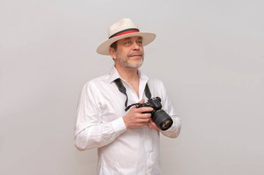 50-55 yaşlarında, şapkalı ve elinde kamerayla beyaz bir arkaplan olan yaşlı bir adam mesafeye bakıyor..