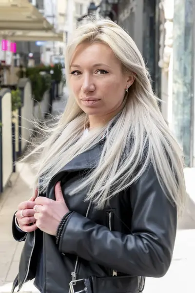 Woman with blonde hair in black jacket standing on blurred city background.