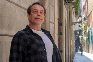 50-55 yaşlarında, dar bir İspanyol sokağında duvara yaslanmış, kollarını göğsüne kavuşturmuş yaşlı bir adamın sokak portresi..