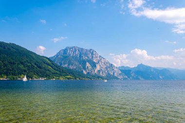 Avusturya 'daki Traunsee Gölü' nün, berrak mavi suların ve arka planda bir sıradağların sergilendiği pitoresk bir manzarası. Göl yelkenlilerle dolu ve gökyüzü birkaç beyaz bulutla dolu parlak bir mavi.. 