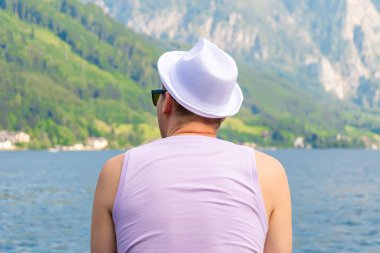 Tişörtlü ve beyaz şapkalı bir adam sırtı kameraya dönük bir şekilde bir dağ manzarasına ve bir dağ gölüne yaslanır..