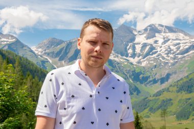 Beyaz gömlekli bir adam yeşil bir dağ yamacının önünde duruyor. Uzakta karla kaplı bir zirvesi var. Güneş parlıyor ve gökyüzü masmavi beyaz bulutlarla.