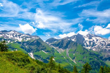 Kısmen karla kaplı, yemyeşil çayırlar ve açık mavi gökyüzü ile çevrili bir dağ manzarası.