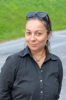 Orta yaşlı, siyah gömlekli ve güneş gözlüklü bir kadının yol kenarına bakıp gülümsemesi..