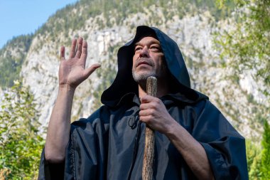 A cloaked monk raises his hand in a dramatic gesture against a backdrop of mountains and greenery during a bright day in nature. clipart