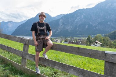 50-55 yaşlarında, geleneksel Tyrolean giysileri içinde, sakin bir kırsal alanda, bir alp zeminine karşı ahşap bir çitin üzerinde oturuyor..