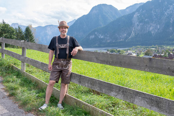 Человек в традиционной тирольской одежде стоит у деревянного забора на фоне горного ландшафта и сельской местности.