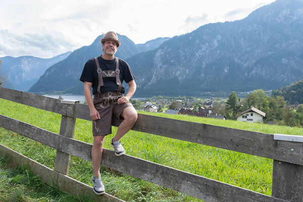 50-55-летний мужчина в традиционной тирольской одежде сидит на деревянном заборе на альпийском фоне в спокойной сельской местности.