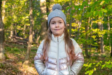 Örgü şapkalı ve ceketli küçük bir kız gündüz vakti ormanda parlak sonbahar yapraklarıyla çevrili gülümsüyor..