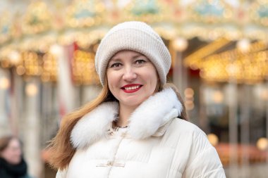 30-35 yaşlarındaki bir kadın, şık beyaz bir ceket ve örgü şapkayla kalabalık bir kış pazarında dikiliyor. Kendine güveni, dekorasyonlarla çevrili şenlikli atmosferde sıcaklık saçıyor..