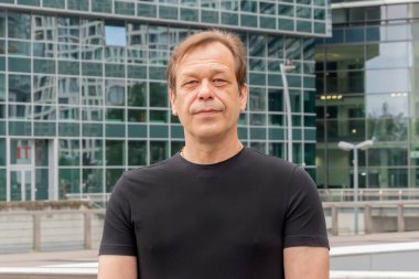 Sağlam vücutlu, kendinden emin yaşlı bir adam cam iş merkezinin arka planında poz veriyor.