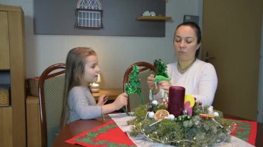 Aile hayatından bir sahne: Anne ve kızı birlikte vakit geçiriyorlar, şenlik dekorasyonu yapıyorlar. Kompozisyonun ortasında bir Noel çelengi ve bir mum var..
