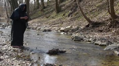 Siyah pelerinli bir keşiş derenin kenarında çıplak ayakla durur ve elleriyle su toplar..