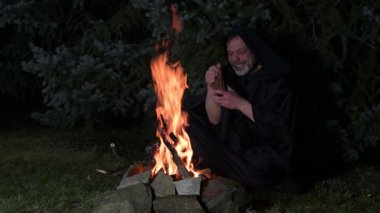 Siyah cüppeli yaşlı bir keşiş gece ormanda ateşin başında otururken cüzdanından para sayıyor.