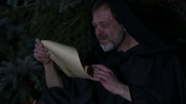 Gizemli bir karakter ormandaki bir ateşin yakınındaki eski bir parşömeni inceliyor.