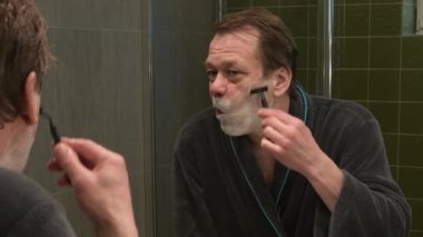 50-55 yaşlarında, sabahlık giymiş yaşlı bir adam banyo aynasının önünde tıraş olurken jilet ve tıraş köpüğü kullanıyor.