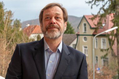 Gri sakallı sağlam bir adam ve Avrupa mimarisinin arka planına karşı dostça bir gülümseme.