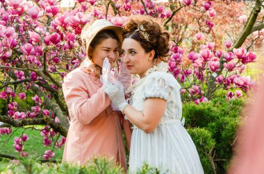 Tarihi kıyafetler içindeki iki kadın, çiçek açan manolyalar karşısında birbirlerine fısıldaşıyorlar.