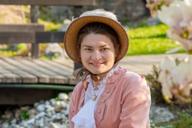 35-40 yaşlarında tarihi Regency elbisesi giymiş bir kadının sokak portresi. Bir bahar parkında çiçek açan ağaçlarla poz veriyor.