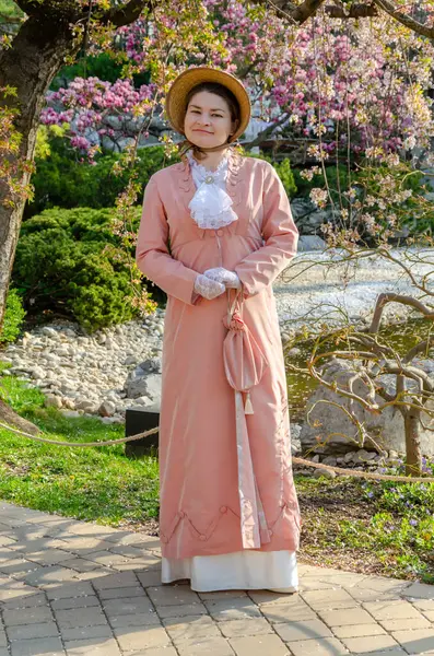 Uzun şeftalili redingot ve şapkalı zarif bir bayan kiraz ağacının altında duruyor.