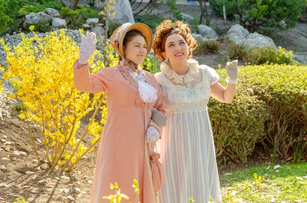 Klasik elbiseli kadınlar çiçek açan bir bahçede sırlarını paylaşırlar.