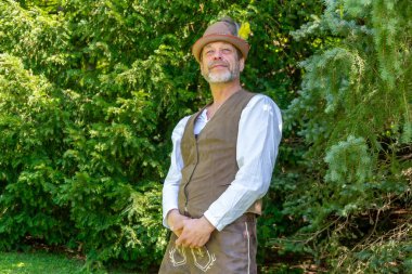 Tyrolean takım elbiseli gülümseyen bir adam kozalaklı bir ağacın yanında duruyor.