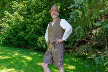 Şapkasında tüy olan bir Tyrolean 'ın halk kıyafeti giymiş bir adam Noel ağaçlarının arka planında duruyor.