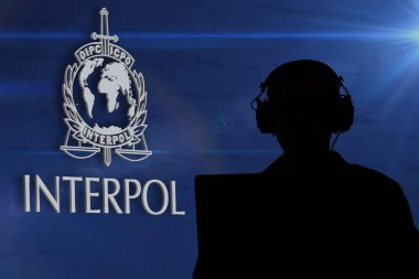 Bilgisayarın önünde kulaklık takan bir adamın silueti Interpol logosunun arka planına karşı siber güvenlik ve gözetlemeyi simgeliyor.
