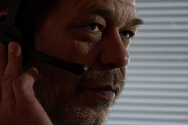 Gözetleme sahnesine yakın çekim. Kulaklıklı bir adamın yüzü perdelerin ardındaki hareketi izliyor.