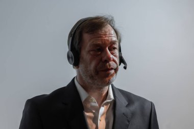 Gri bir duvara karşı mikrofonlu takım elbiseli bir adam. Ciddi bir ifadeyle orta yaşlı bir adam, takım elbise giyiyor, kulaklıkla konuşuyor ya da dinliyor.