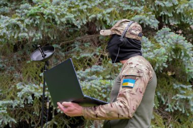 Ukrayna bayrağı taşıyan kamuflajlı bir asker ormanda radyo keşfi yapan bir mikrofon ve dizüstü bilgisayar kullanıyor.