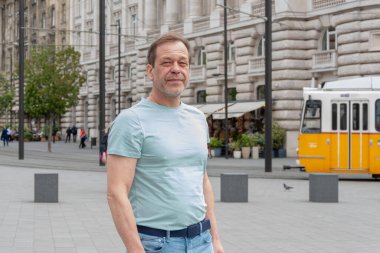 50-55 yaşlarındaki yetişkin bir adamın şehir sokaklarındaki portresi. Açık renkli tişörtlü bir adam klasik Avrupa mimarisinin ve sarı tramvayın arka planına karşı duruyor.