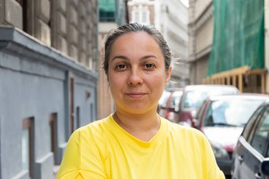 Şehir caddesinde duran parlak sarı tişörtlü orta yaşlı bir kadının portresi. Yüze odaklanmış, arka plan bulanık, binalar ve arabalar görünür, kentsel bir atmosfer yaratıyor.