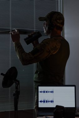 Taktik üniformalı bir memur, bir laptop ve mikrofonun yanında dururken dürbün kullanır. Operasyonel çalışma atmosferi