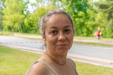 Tarafsız bir ifadeyle orta yaşlı bir kadın, yaz günü yeşil ağaçların ve yolların arkasında poz veriyor.