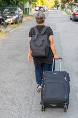 Sırt çantası ve bavulu olan bir kadın sakin bir caddede yürüyor.