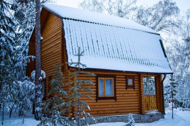 Karla kaplı ahşap kulübe, karlı bir ormanda, buzlu pencereleri ve uzun ağaçlarla çevrili, sıcacık bir kış inzivası.