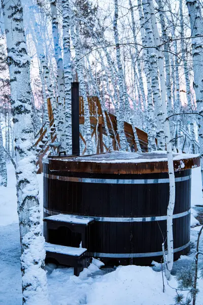 A wooden hot tub is nestled in a snowy forest, surrounded by icy icicles and bare tree trunks. The winter scene is serene, with electric blue accents reflecting off the glass
