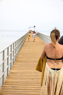 Renkli bir bikini giyen bir kadın, güneşli, ahşap bir iskelede rahatça yürüyor.